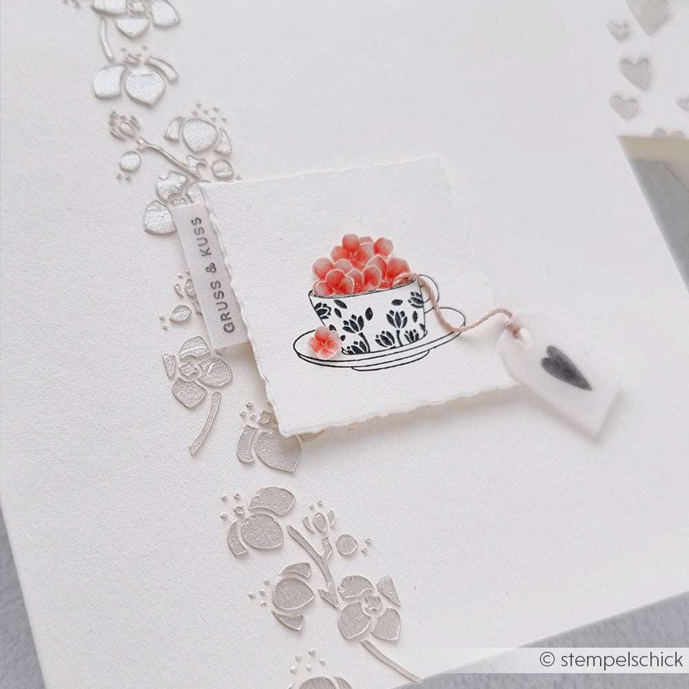 Handgefertigte Karte von Alexandra Renke mit geprägten Blumenmustern und einer kleinen geblümten Tasse, gefüllt mit rosa Herzen - perfekt, um süße Momente zu teilen.