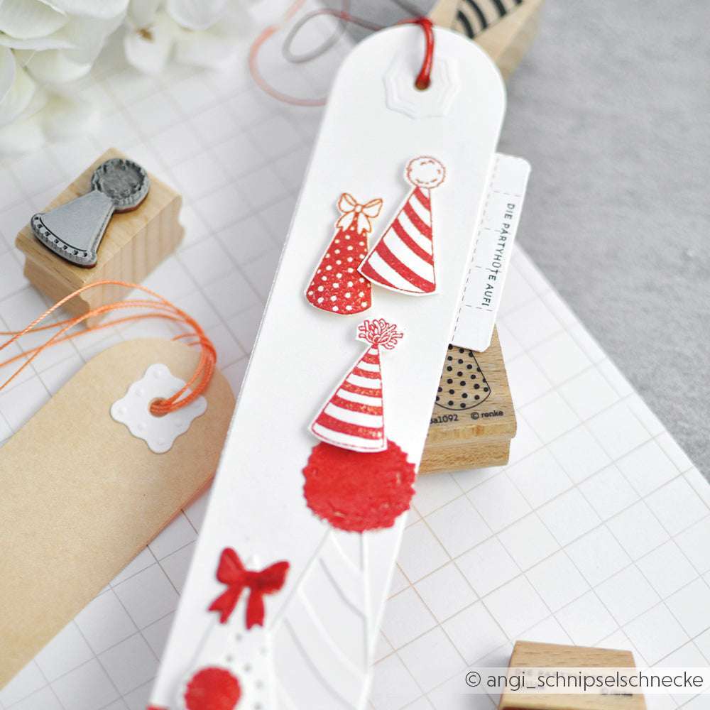 Ein weißes, handgefertigtes Lesezeichen von Alexandra Renke mit roter Partyhütchen-Dekoration, Schleife und Holzstempel-Prägung, dargestellt auf einem Gitter-Hintergrund.