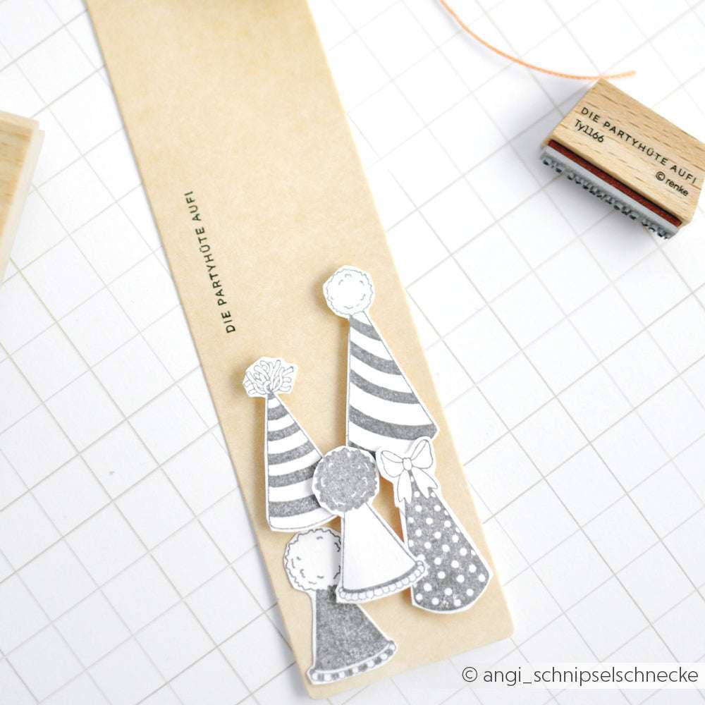 Fünf schwarz-weiße Partyhüte aus Papier zieren ein Lesezeichen von Alexandra Renke, daneben ein Partyhütchen-Stempel und eine Schnur, alles vor einem gitterförmigen Hintergrund.