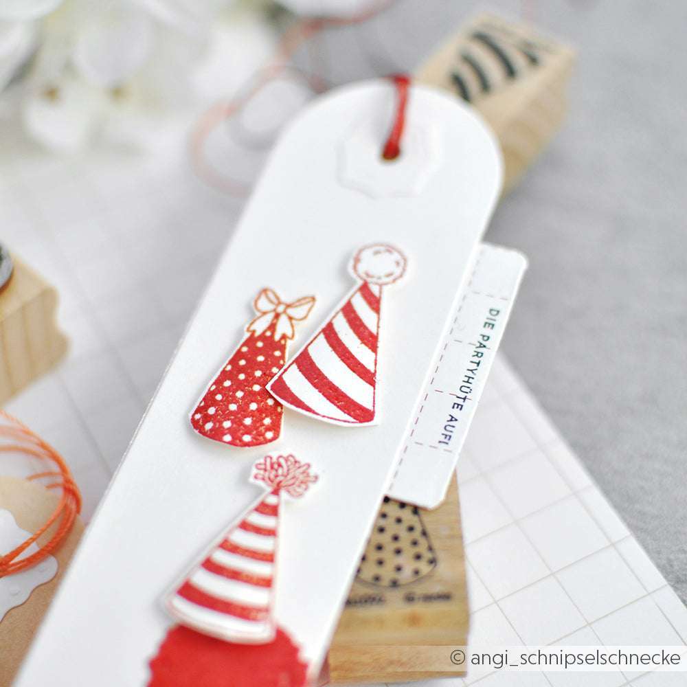 Nahaufnahme eines handgefertigten Lesezeichens von Alexandra Renke mit drei roten und weißen Partyhütchen, mit Schnur und Papierakzenten - perfekt, um Ihren Seiten ein festliches Flair zu verleihen.