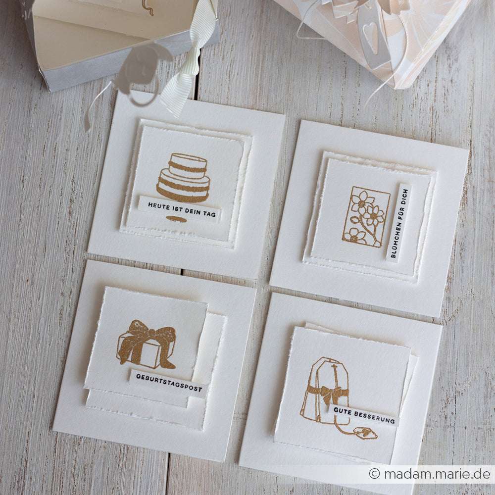 Vier quadratische Grußkarten von Alexandra Renke mit detaillierten Motiven und goldenen Illustrationen für verschiedene Anlässe sind auf einer hellen Holzoberfläche angeordnet.