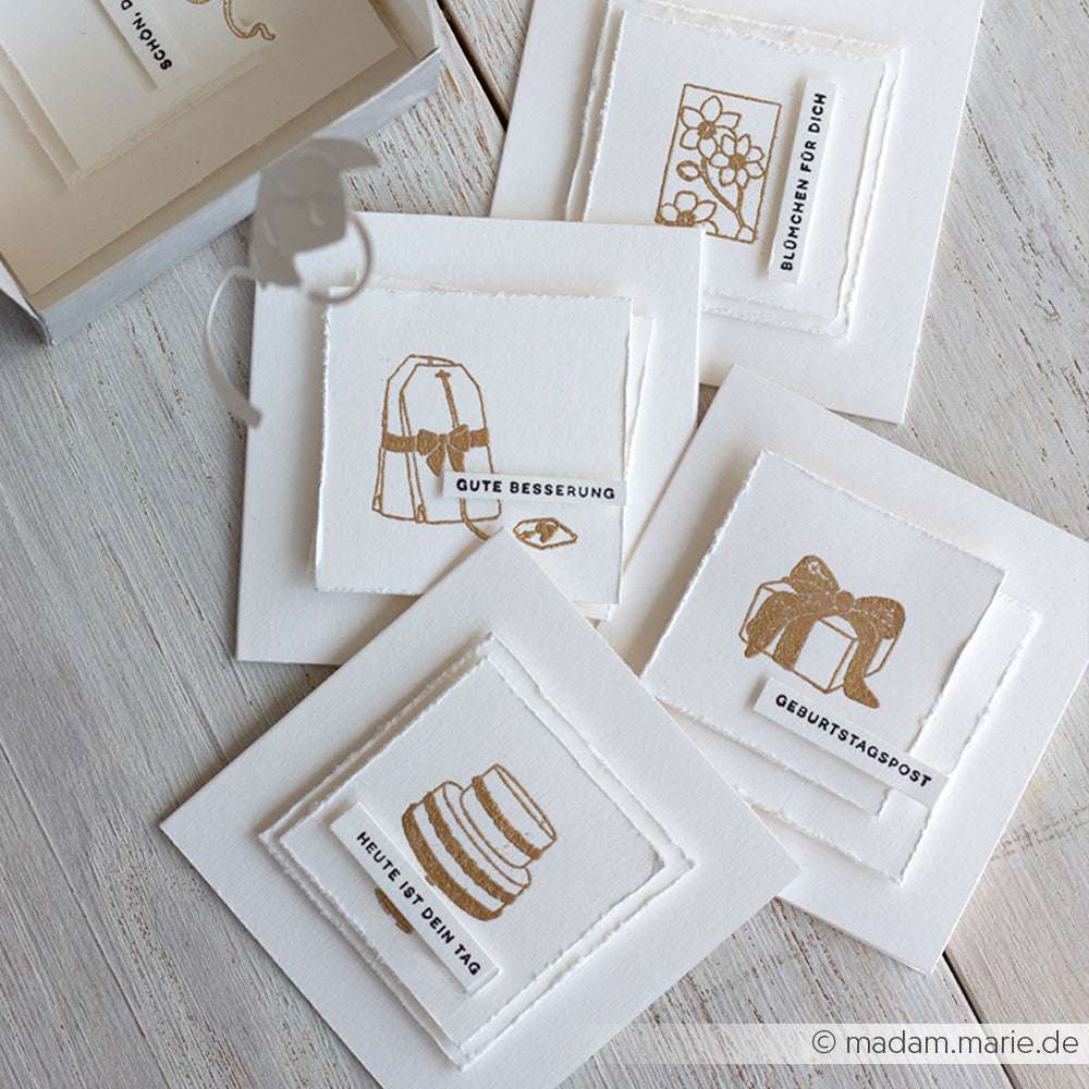 Vier weiße Karten mit minimalistischen goldenen Stempelmotiven und deutschen Sprüchen von Alexandra Renke, arrangiert auf einer hellen Holzoberfläche - die perfekte Inspiration für Geschenkstempel- oder Holzstempel-Fans.