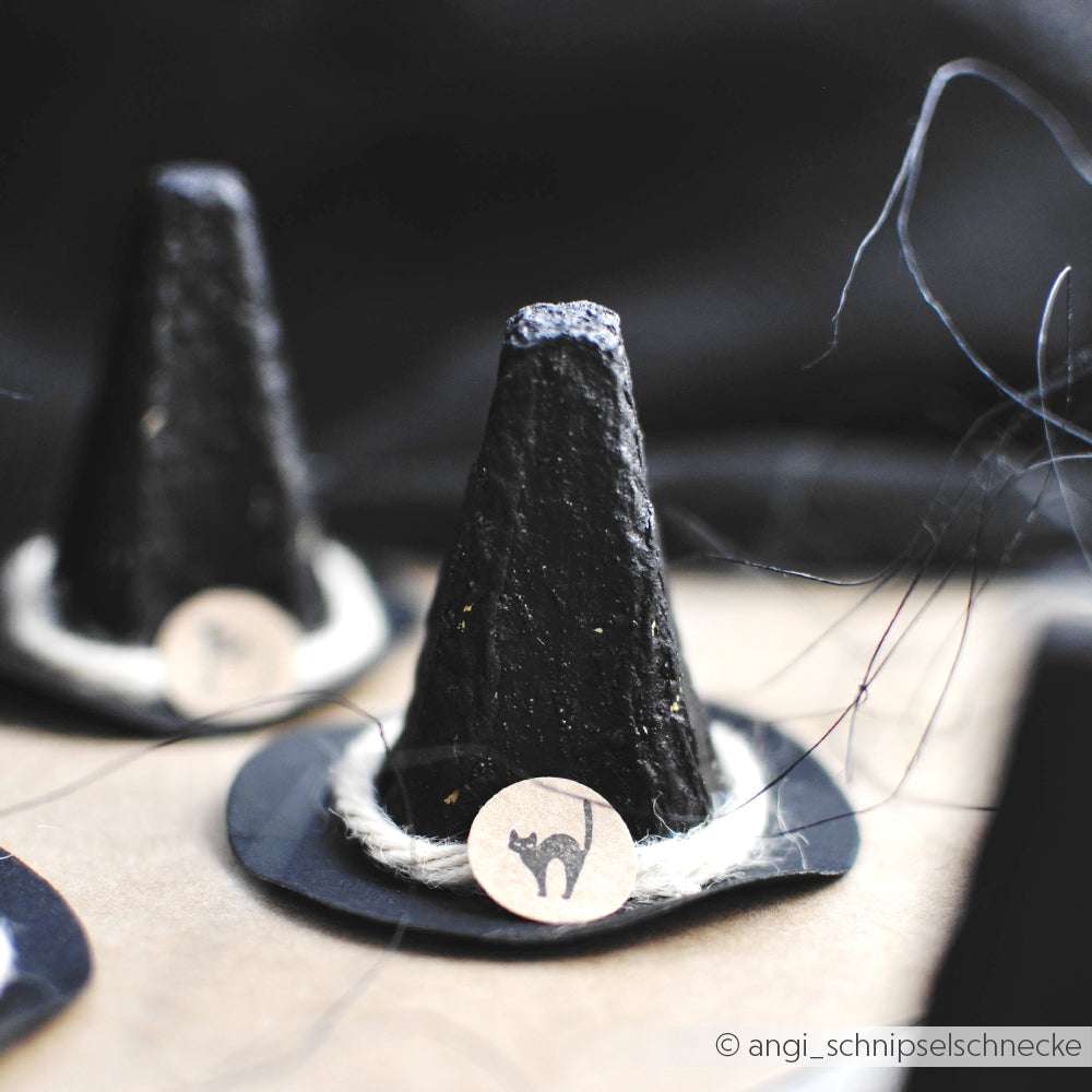 Kleiner schwarzer Hexenhut mit einem Mini Schwarze Katze Anhänger, der auf einer braunen Oberfläche sitzt - ideal zum Kombinieren mit Alexandra Renke Akzent-Stempel oder Holzstempel in Ihren Bastelprojekten.