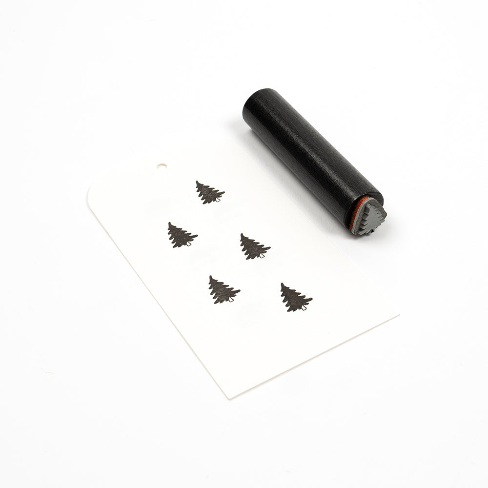 Ein kleiner zylindrischer Holzstempel und ein Papieranhänger mit fünf aufgestempelten Symbolen für immergrüne Bäume.