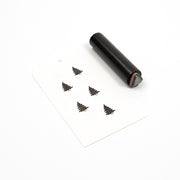 Ein kleiner zylindrischer Holzstempel und ein Papieranhänger mit fünf aufgestempelten Symbolen für immergrüne Bäume.