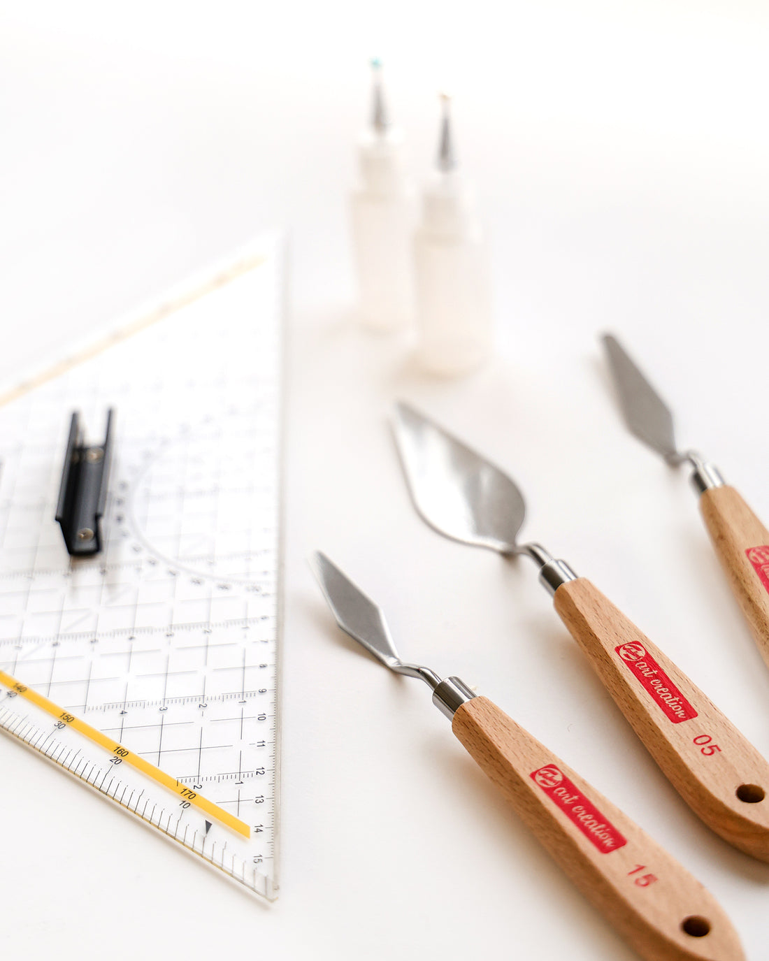 Drei Spachtel, ein Lineal, ein Stift und zwei Flaschen auf einer weißen Fläche.