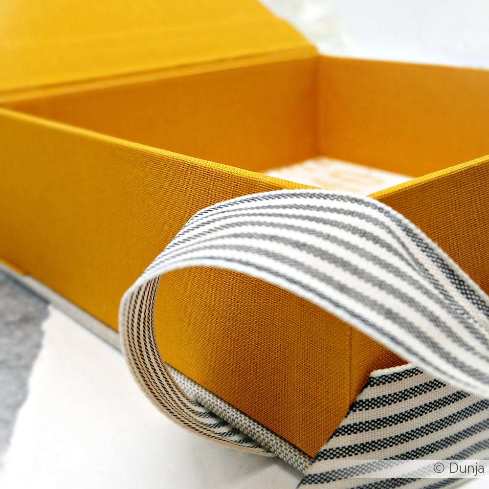 Eine gelbe ☆ Schachtel mit einem schwarz-weiß gestreiften Band, das teilweise um die Schachtel gewickelt ist und mit einem Hauch von Buchleinen für Eleganz sorgt.