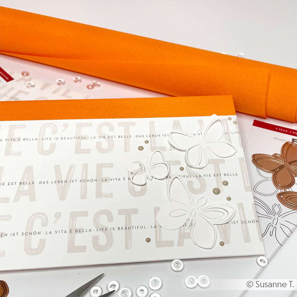 Eine ☆ handgefertigte Karte mit geprägten Schmetterlingen und zweisprachigem Text, eingebettet in Bastelmaterial, orangefarbenem Papier und elegantem Buchleinen für einen raffinierten Touch.