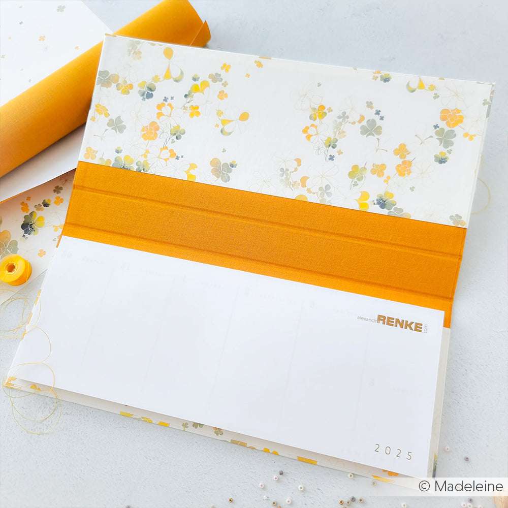 Der ☆ Kalender ist mit einem floralen Muster und einem orangefarbenen Band versehen, aus elegantem Buchleinen gefertigt und mit einer leeren Seite 2025 auf einer weißen Oberfläche aufgeschlagen.