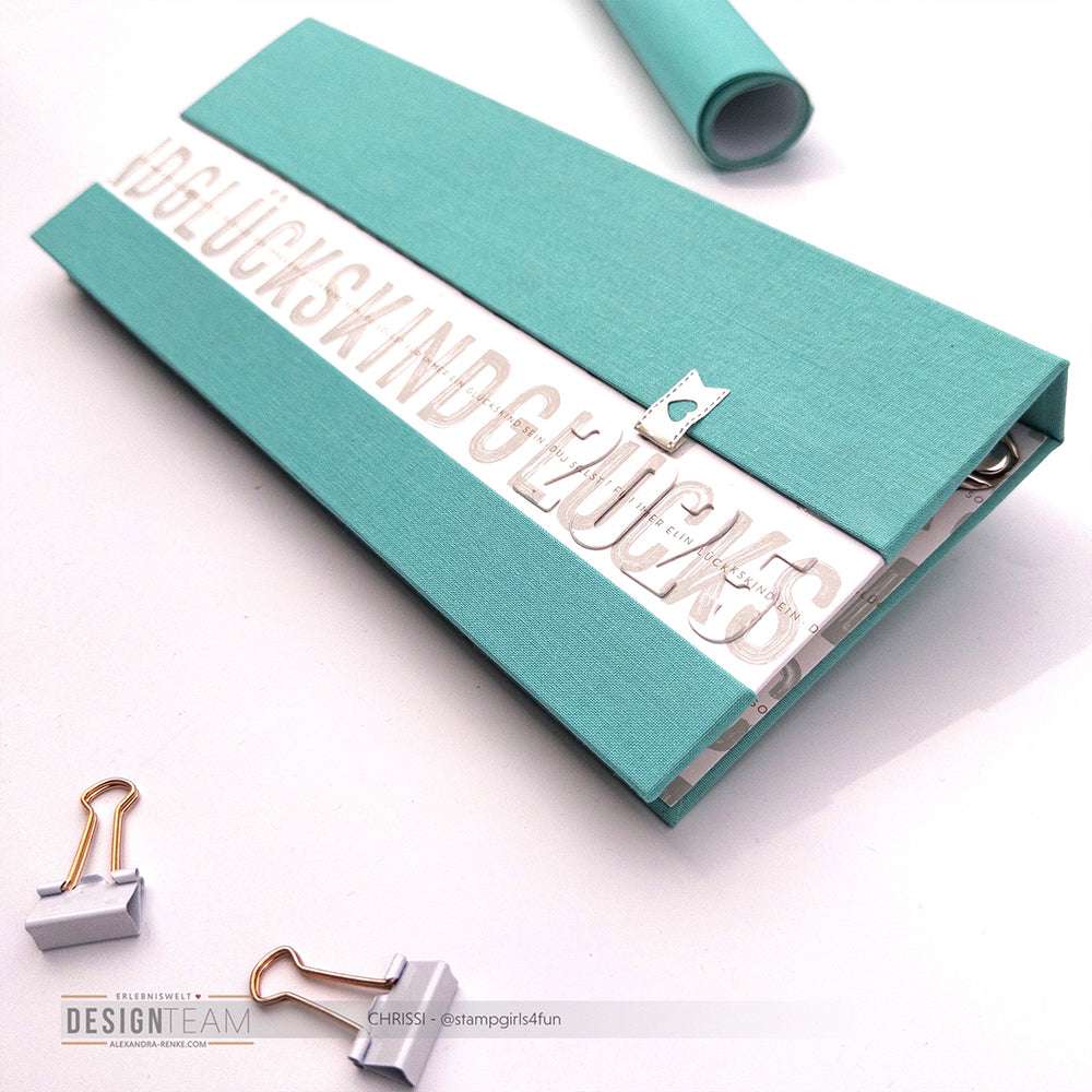 Eine tealfarbene Buchleinen-Schreibmappe von ☆ mit dem Text "Glückskind", mit Heftklammern und einem sichtbaren Blatt Papier im Inneren.