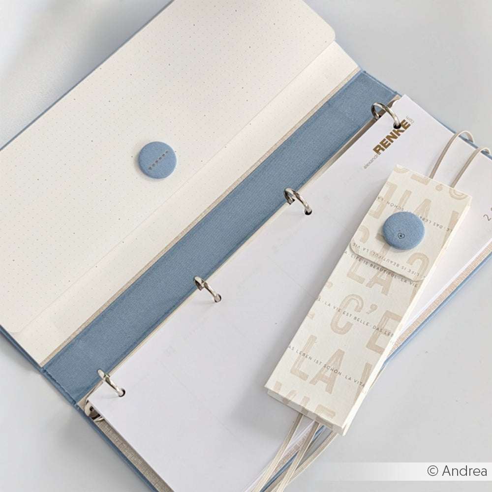 Offene blau-weiße ☆ Mappe mit gepunktetem Papier und einem knopfverzierten Lesezeichen mit Buchleinen-Akzenten auf weißer Unterlage.