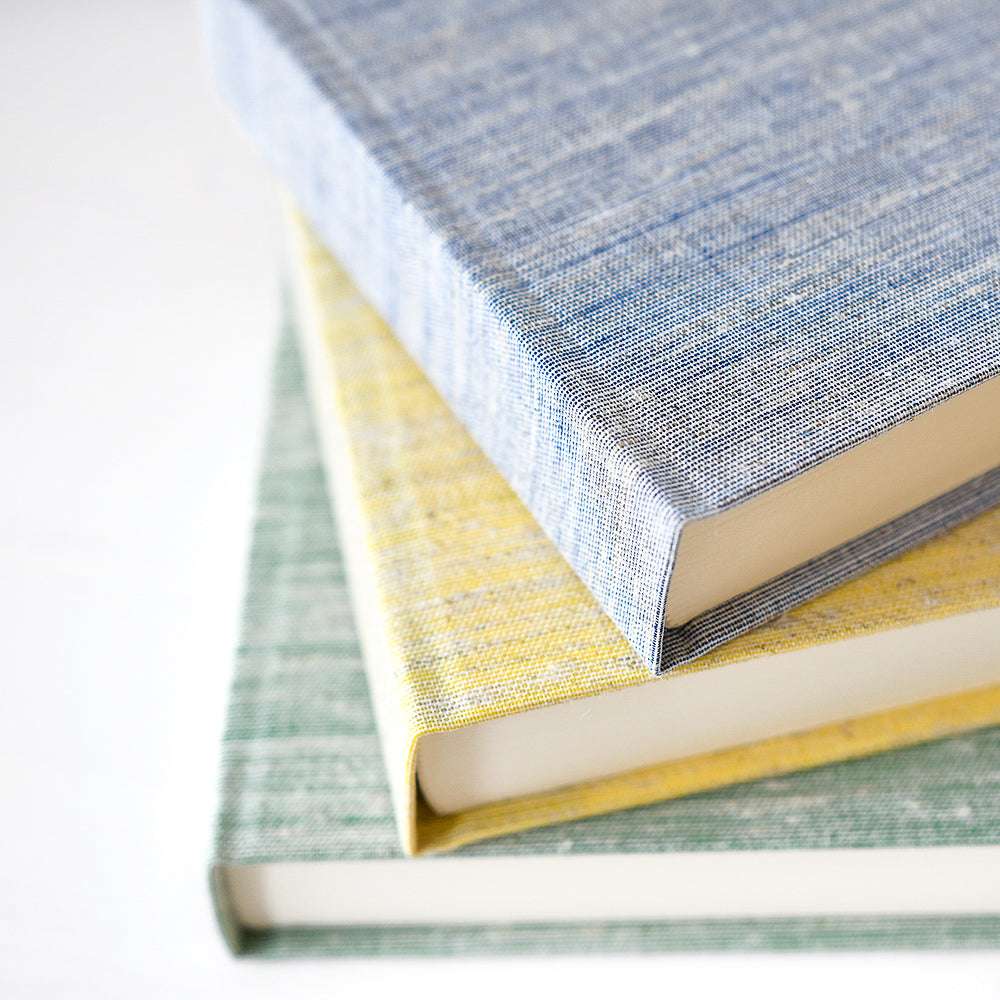 Drei ☆ Hardcover-Bücher, die auf einer weißen Fläche gestapelt sind, mit strukturierten Buchleineneinbänden in Blau, Gelb und Grün.