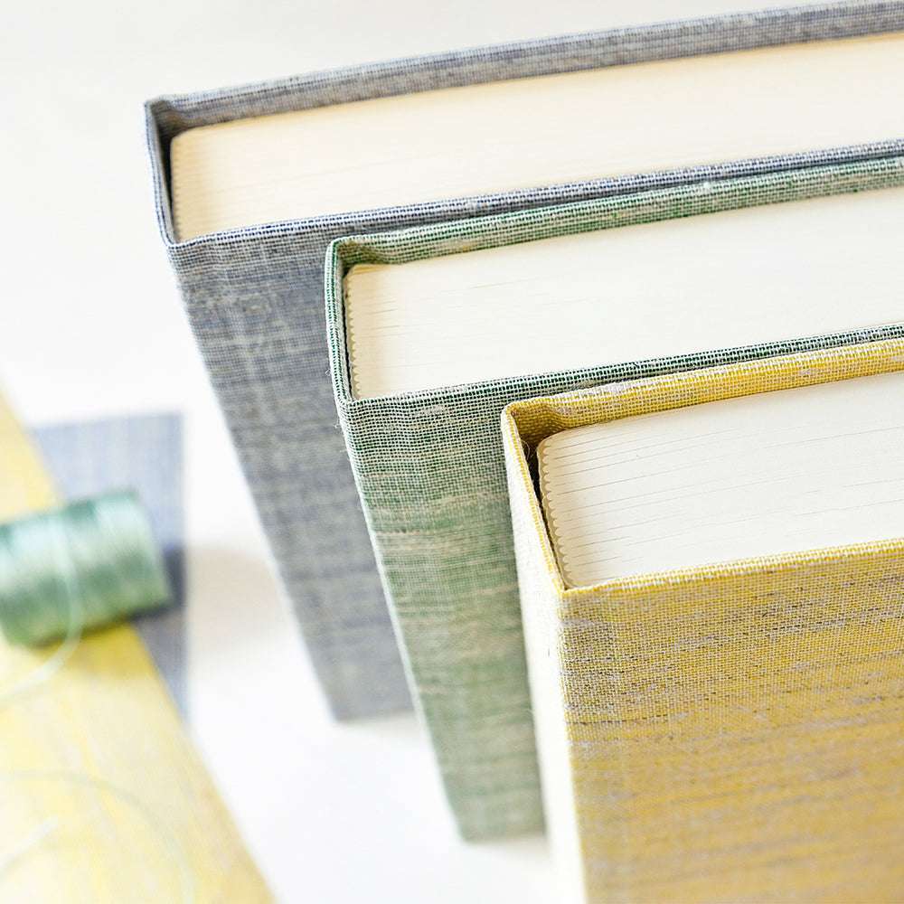 Drei ☆ Hardcover-Bücher mit strukturierten Buchleineneinbänden in Grau, Grün und Gelb stehen neben einer Spule mit grünem Garn.