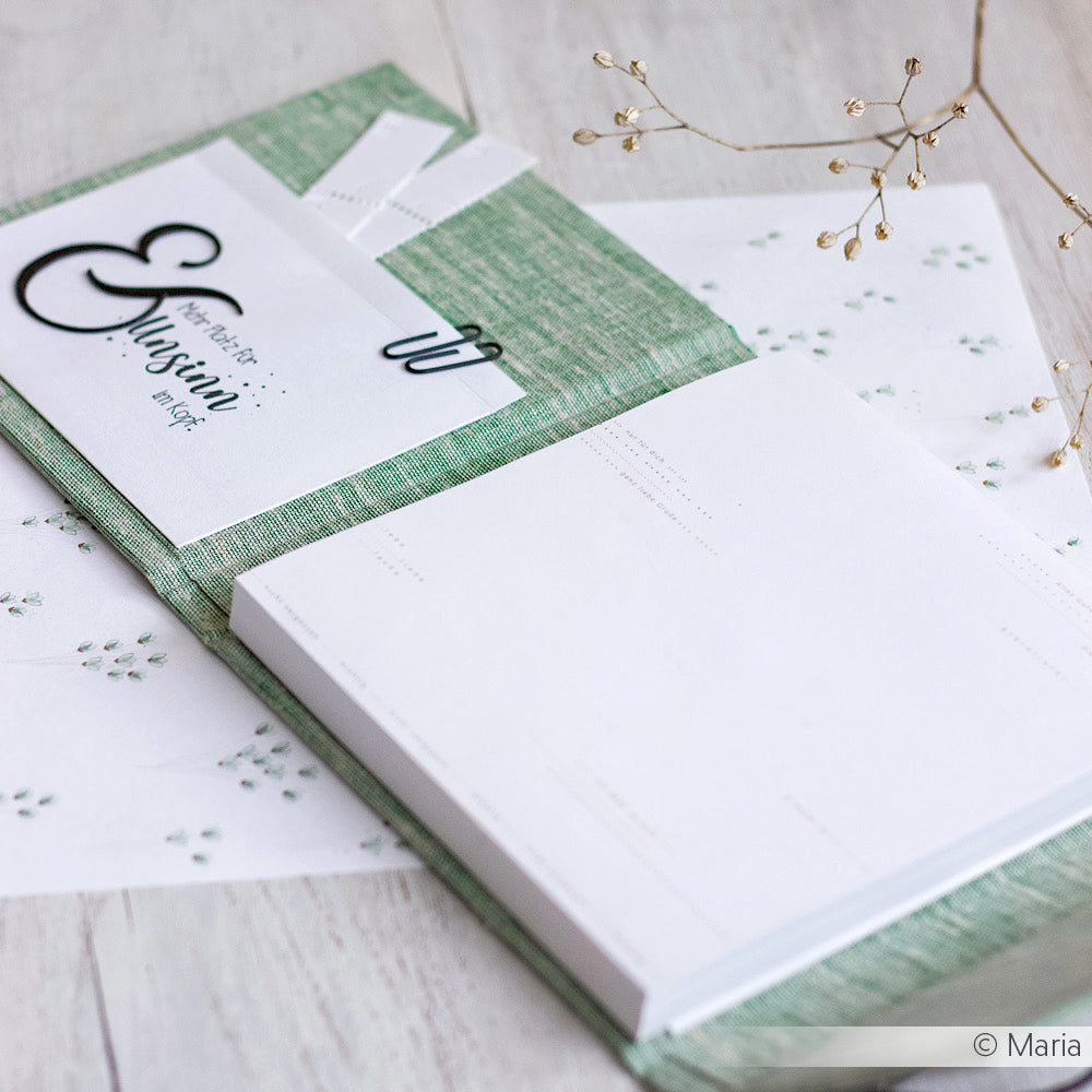 Aufgeschlagenes grünes Notizbuch aus Buchleinen mit leeren Seiten, begleitet von weißen Karten und zartem Papier mit botanischen Motiven, ausgestellt auf einer hellen Fläche.