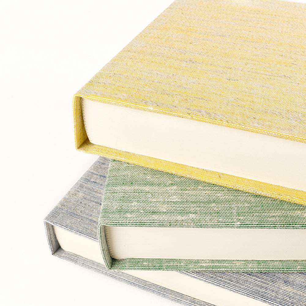 Drei gestapelte Hardcover-Bücher, jeweils in gelbes, grünes und blaues Buchleinen-Naturgewebe aus ☆ eingewickelt, zeigen ihre strukturierten Einbände auf weißem Hintergrund - perfekt, um die im Offsetdruck verwendeten hochwertigen Materialien hervorzuheben.