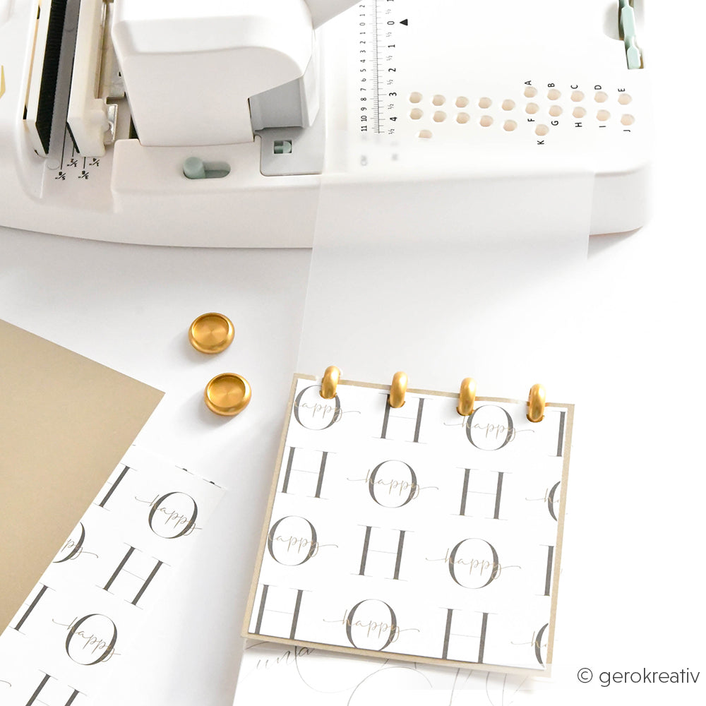 Ein Papierlocher, goldene Bindescheiben und ein Notizblock mit Monogramm von Alexandra Renke sind auf einer weißen Oberfläche angeordnet - ideal, um Ihr eigenes Mini-Album zu gestalten oder mit einzigartigen Bindeelementen zu personalisieren.