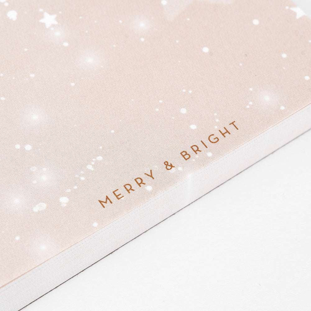 Nahaufnahme eines Alexandra Renke Notizblocks mit der Aufschrift "MERRY & BRIGHT" in goldenen Buchstaben auf einem hellen, an den Sternenhimmel angelehnten Hintergrund.
