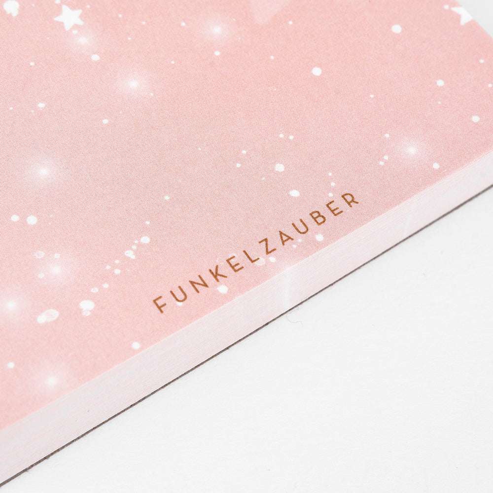 Der rosafarbene Notizblock von Alexandra Renke ist mit weißen, sternförmigen Glitzern verziert, die an den Sternenhimmel Funkelzauber erinnern. Am unteren Rand ist der Schriftzug FUNKELZAUBER" in Gold aufgedruckt.