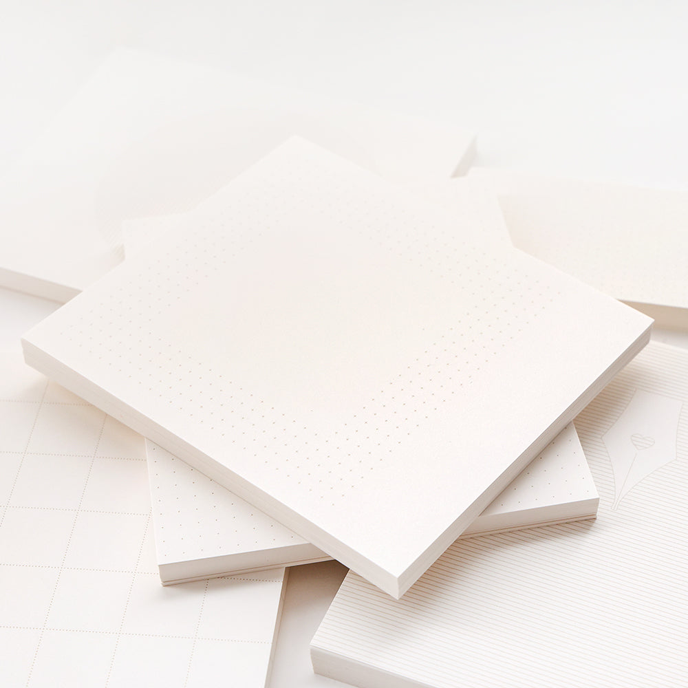 Ein Stapel leerer Notizblockseiten von Alexandra Renke mit subtilen Raster- und Punktmustern auf weißem Papier, ideal für Bullet-Journal-Setups, arrangiert in weichem Licht.