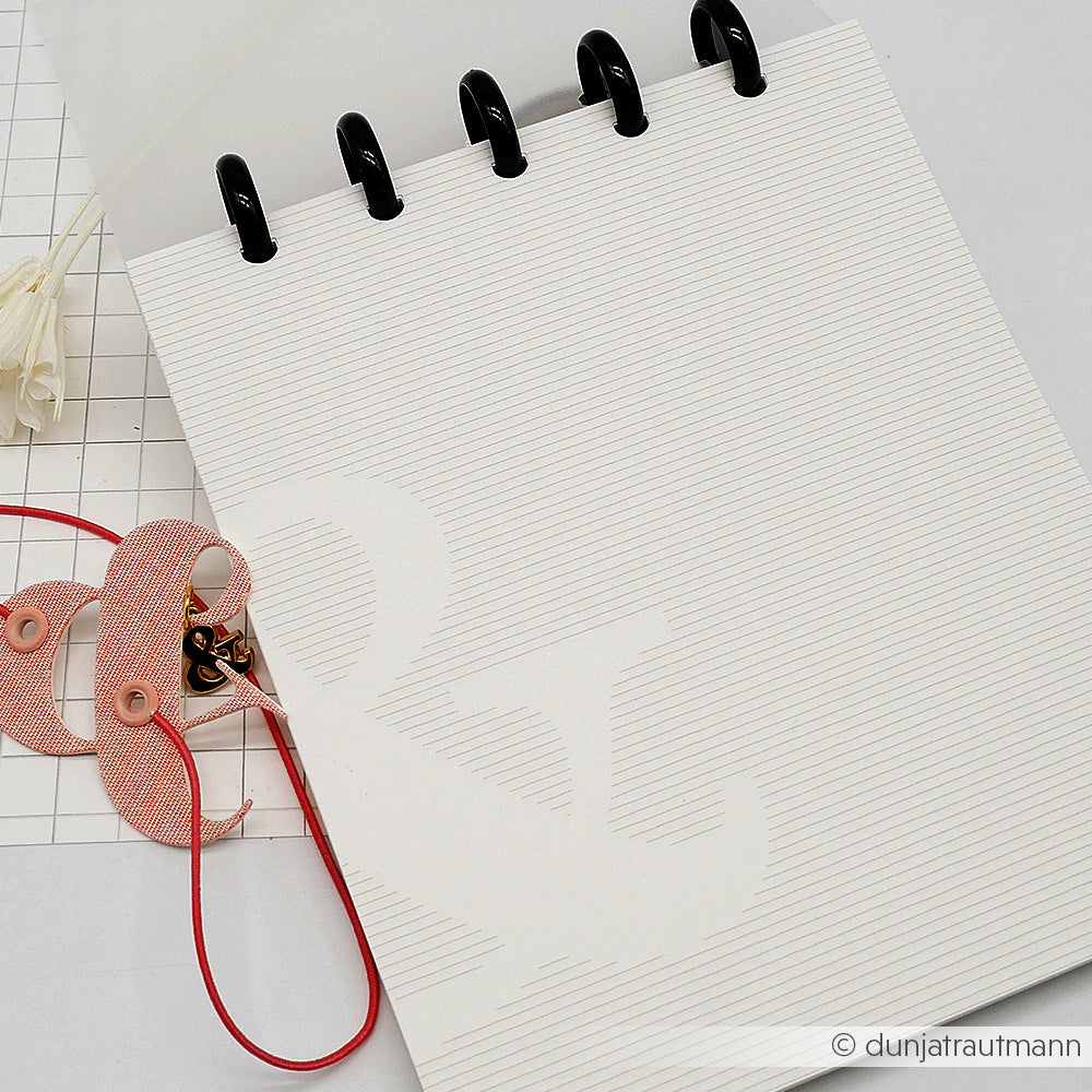 Ein Alexandra Renke Notizblock mit linierten Seiten liegt aufgeschlagen neben rosa Knöpfen und roter Schnur auf einer weißen Fläche, perfekt für Ihr nächstes Bullet-Journal-Projekt.