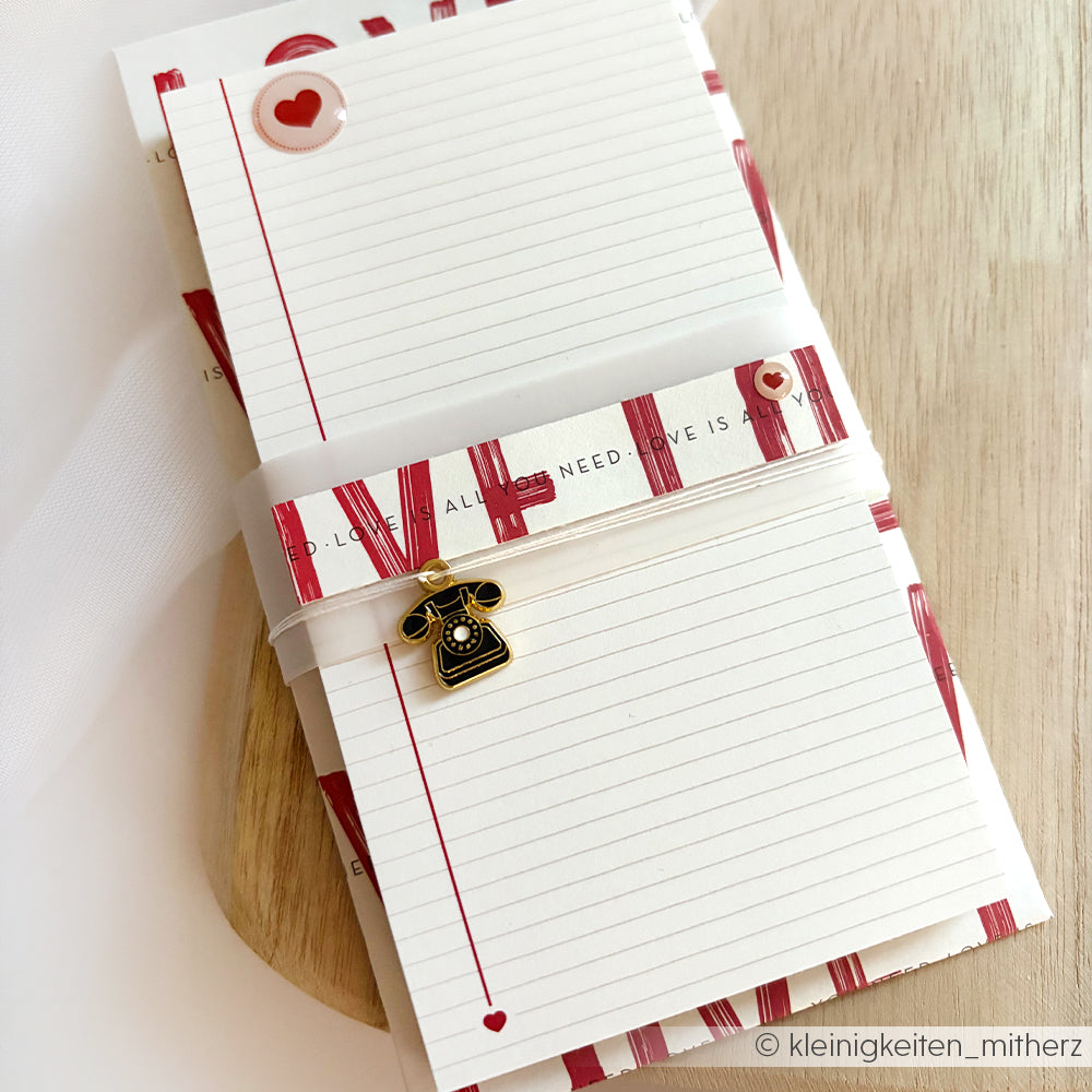 Ein Alexandra Renke Notizblock mit roten Streifen, Herzmotiven und einem Vintage-Telefon-Charme ruht auf einer hölzernen Oberfläche - perfekt für Bullet Journal-Fans oder zum Notieren auf losen Blättern.