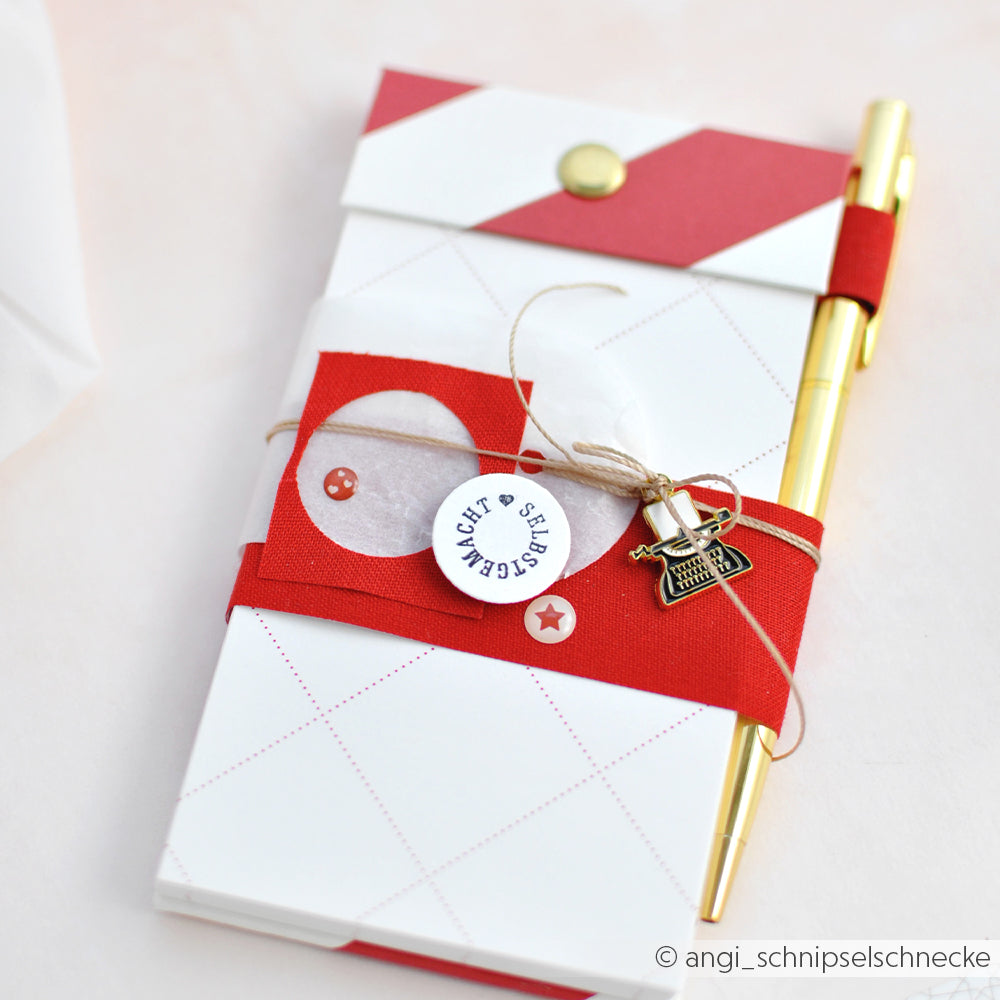Ein goldenes Stift- und Notizblockset von Alexandra Renke, verpackt in rotem Band mit einem Mini-Schreibmaschinenanhänger und einem dekorativen Anhänger - perfekt für Bullet-Journal-Fans.