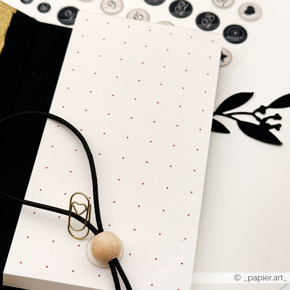 Ein gepunkteter Bullet-Journal Notizblock von Alexandra Renke mit einer schwarzen Kordel, einer Holzperle, einer herzförmigen Büroklammer und schwarz-weißen Aufklebern.