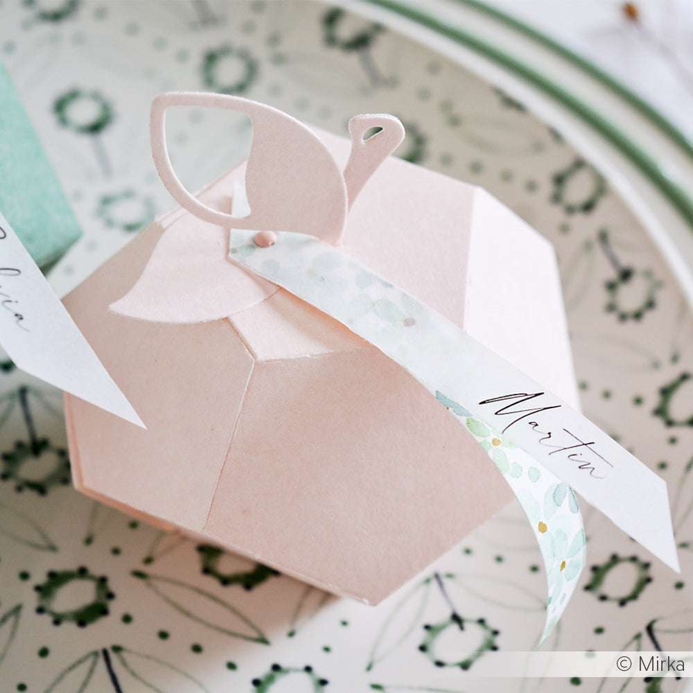 Zartrosa, geometrische Geschenkbox von Alexandra Renke mit Schwan-Topper und Namensschild auf einer gemusterten Platte. Verziert mit 3mm Rosa Brads für einen charmanten Scrapbooking-Akzent.