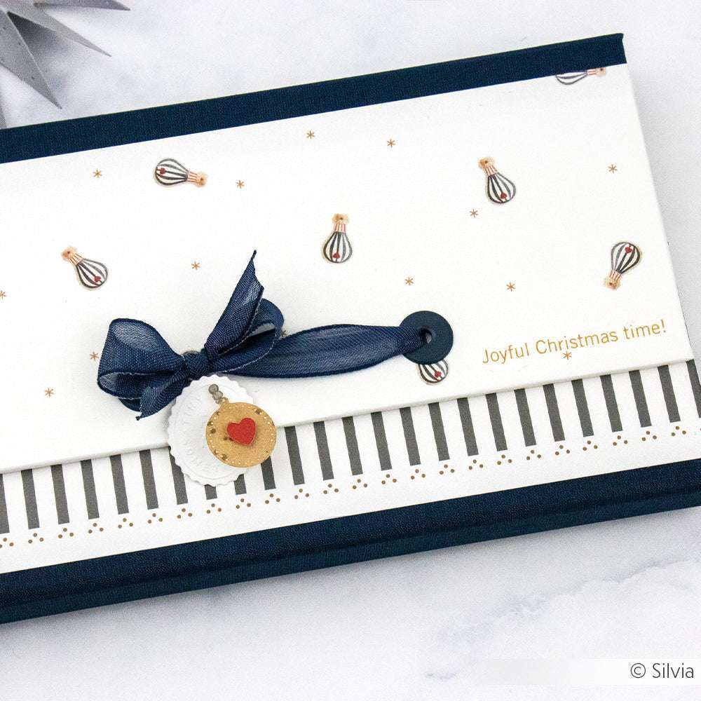 Weißer Umschlag mit kosmosblauem Band, festlichen Anhängern und dem Text "Joyful Christmas time!" auf einer gestreiften Schachtel von Alexandra Renke - ideal zum Buchbinden oder Verschenken.