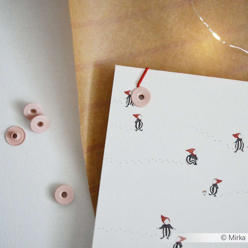 Weiße Karte von Alexandra Renke mit kleinen Figuren mit roten Hüten, rotem Band, rosa Knöpfen und braunem Papierhintergrund - ideal als dekoratives Element für Buchbindeprojekte.