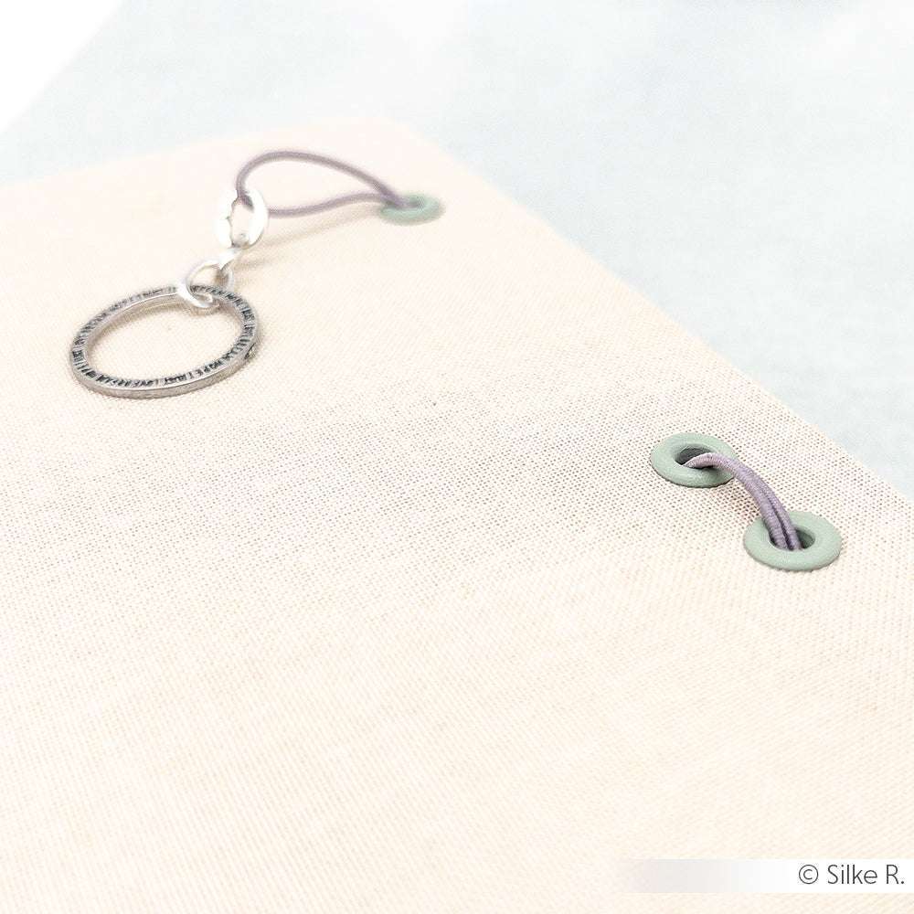 Nahaufnahme eines beigen Zeitschrifteneinbands aus Stoff von Alexandra Renke mit Buchbinden, Ösen mit Unterlegscheibe und einer lilafarbenen, durchgefädelten Gummischlaufe.