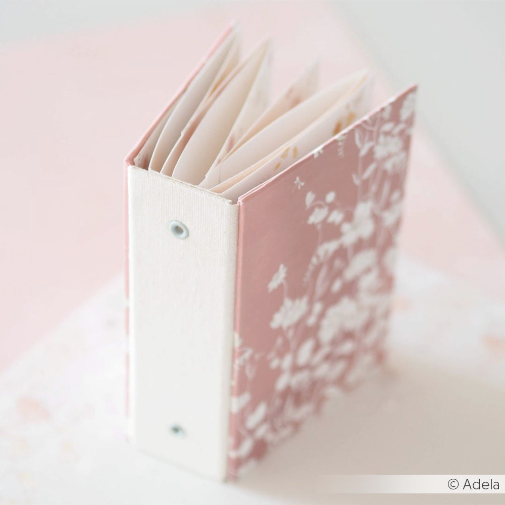 Ein kleines, rosafarbenes, floral gemustertes Minialben von Alexandra Renke mit weißem Rücken steht offen auf einem hellen Hintergrund - perfekt für Buchbinden-Projekte oder kreatives Journaling.