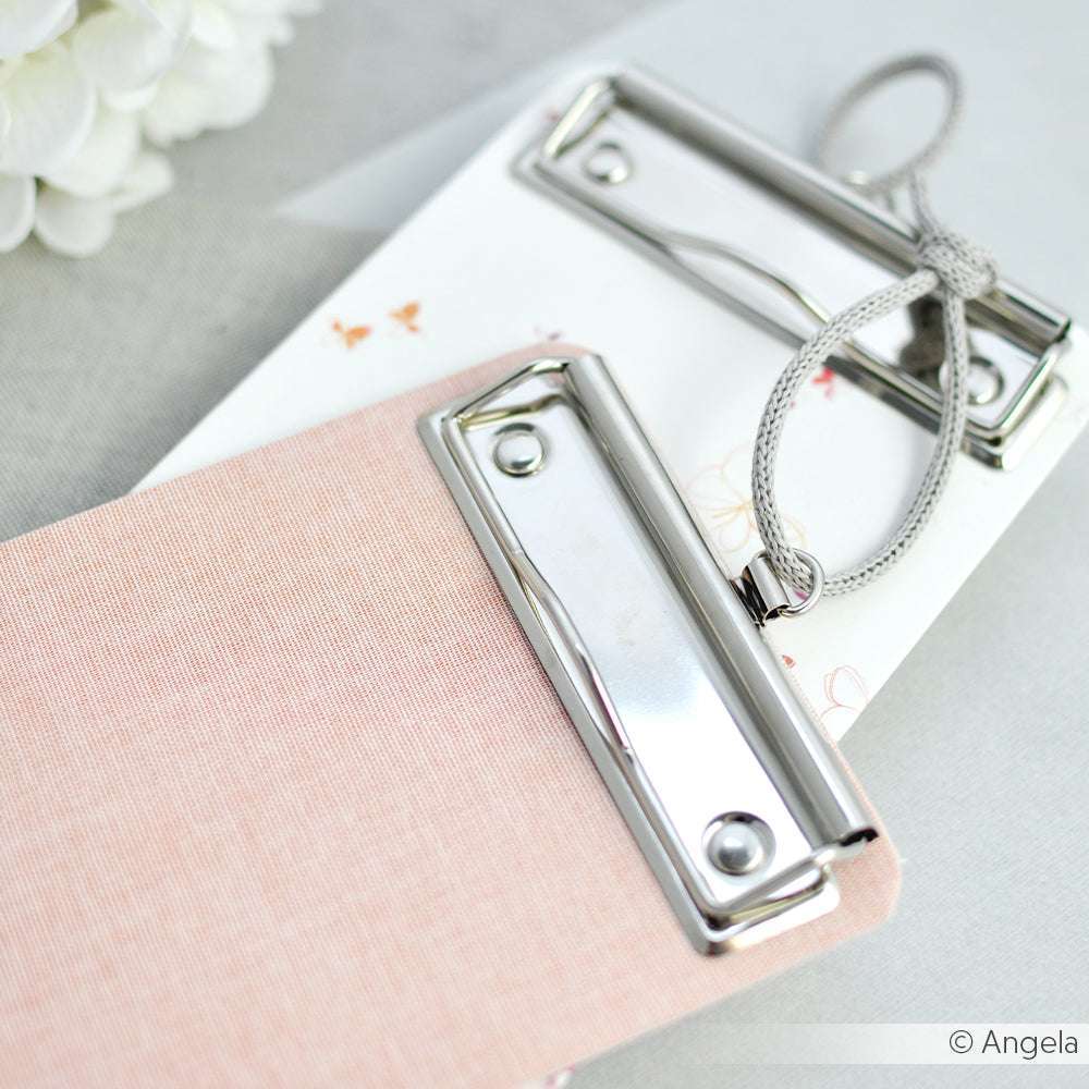 Zwei kleine ☆ Klemmbretter, ein rosa und ein weißes, jeweils mit einem stabilen Metallklammermechanismus, liegen auf einer hellen Fläche.