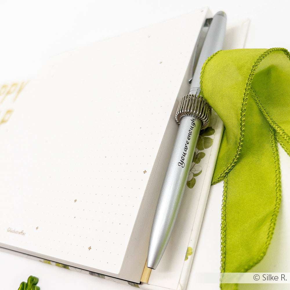 Ein ☆ silberner Stift mit der Aufschrift "Du bist genug" ruht auf einem aufgeschlagenen gepunkteten Notizbuch, daneben ein grünes Band und ein praktischer Stifthalter selbstklebend.