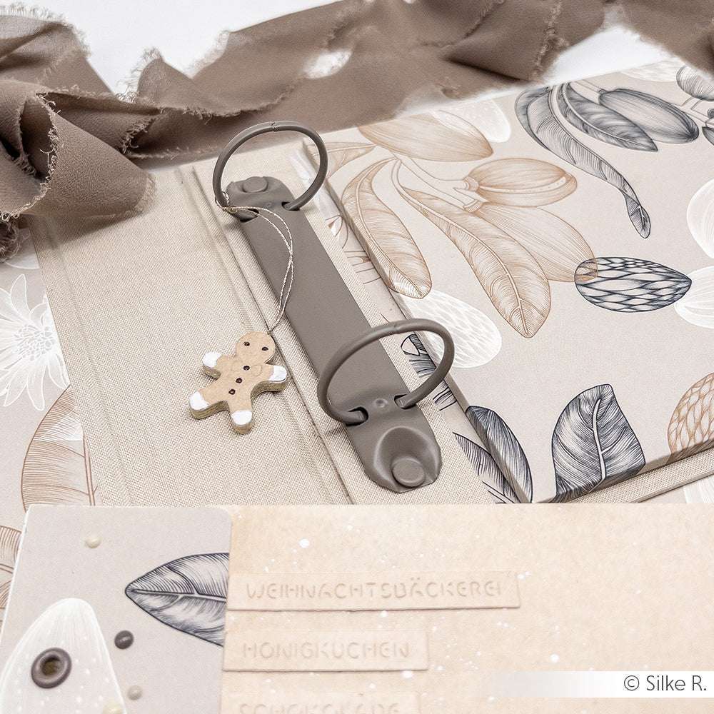 Ein beigefarbener Binder von Alexandra Renke mit Blattmotiven und einem Lebkuchenmann-Ornament liegt auf Stoff - ideal für Buchbindeprojekte oder zum Basteln charmanter Mini-Alben.
