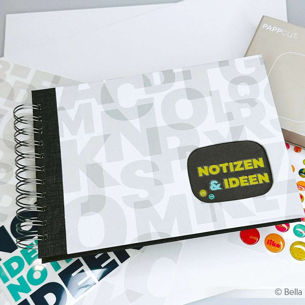 Das Spiralnotizbuch Notizen & Ideen von Alexandra Renke liegt auf einem Schreibtisch mit bunten Stickern, Bucheinbandpapier und Briefpapier - ideal für Buchbinden-Projekte und kreative Notizen.