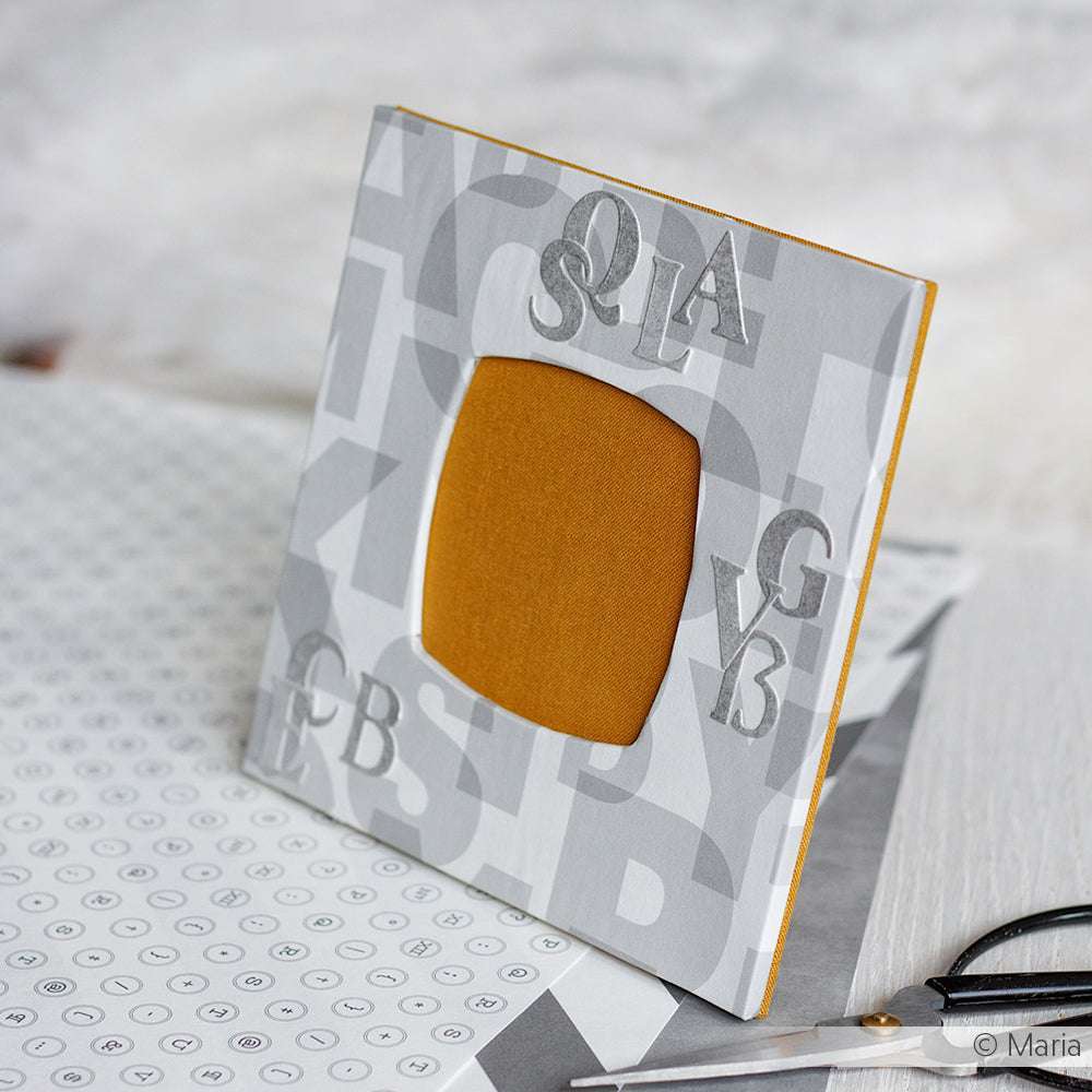 Der grau-weiße Bilderrahmen von Alexandra Renke mit verstreuten Buchstaben und einem leeren senfgelben Platz in der Mitte ist aus einzigartigem Designpapier gefertigt - perfekt für kreative Buchbindeprojekte.