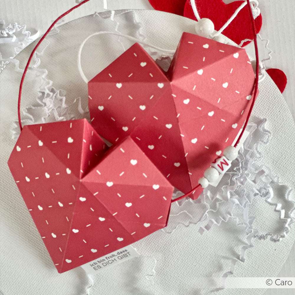 Zwei Origami-Papierherzen mit weißen Punkten von Alexandra Renke Premiumrot liegen auf einer Spitze, die mit Perlen und einem Anhänger verziert ist - die perfekte Inspiration für kreative Buchbindearbeiten oder einzigartige Buchumschläge.