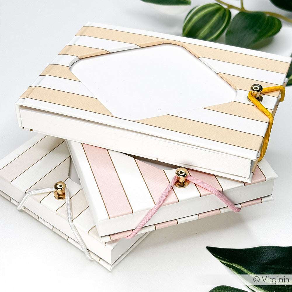 Drei gestreifte, rechteckige Schachteln von Alexandra Renke, eingewickelt in Bucheinbandpapier mit Herzbordüre mit Streifen Gelb und elastischen Verschlüssen, sind auf einer weißen Fläche mit grünen Blättern gestapelt - eine Anspielung auf traditionelle Buchbinden.