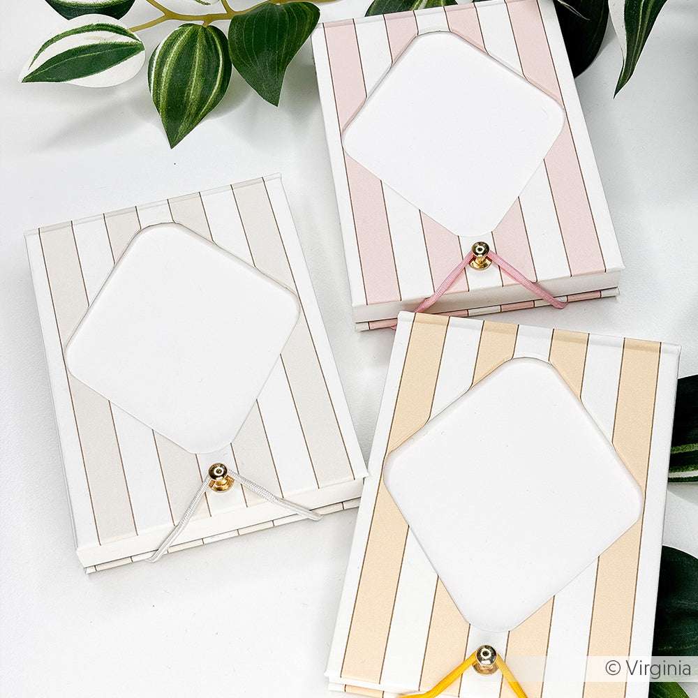 Drei gestreifte Notizblöcke von Alexandra Renke mit Gold- und Bandakzenten, hergestellt aus edle Designpapiere und Bucheinbandpapier, werden auf einer weißen Fläche mit grünen Blättern in der Nähe präsentiert.