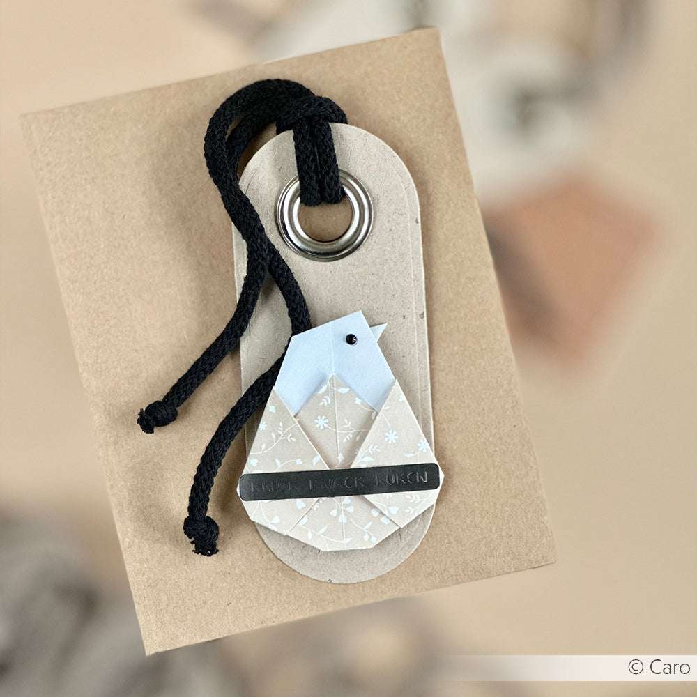 Eine Geschenkbox von Alexandra Renke mit einem vogelförmigen Origami-Anhänger aus Designpapier und einer schwarzen Kordel, die durch eine Metallöse gefädelt ist - ideal für kreatives Buchbinden.