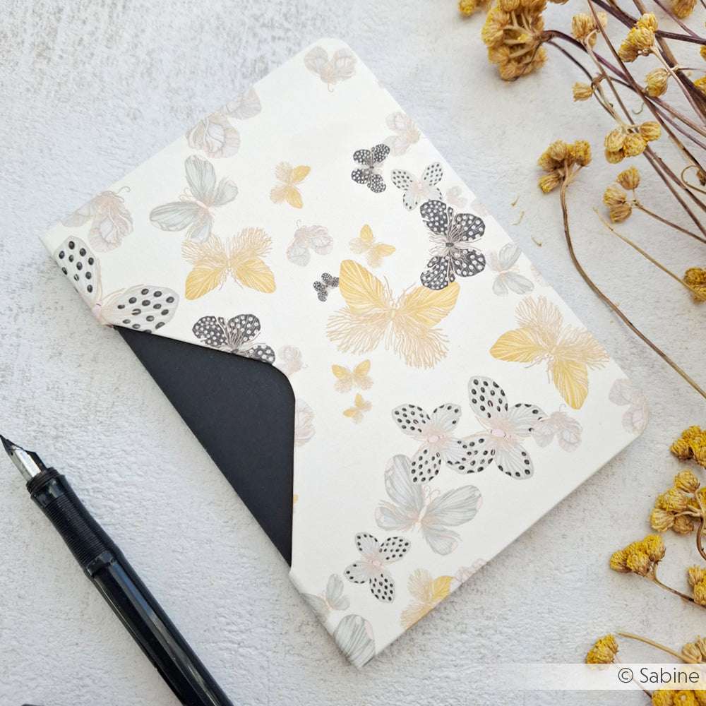 Ein Umschlag mit Blumenmuster von Alexandra Renke Designpapier hält eine schwarze Karte, die neben einem Stift und getrockneten gelben Blumen auf einer hellen Fläche zu sehen ist.