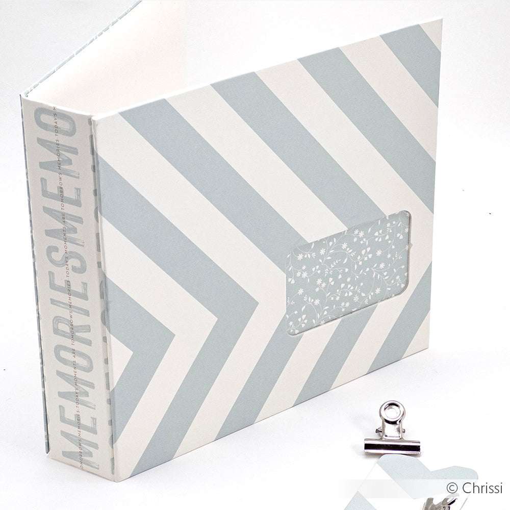 Ein hellblau-weißes Fotoalbum von Alexandra Renke mit dem Schriftzug MEMORIES" auf dem Buchrücken aus Blockstreifen Bleu Bucheinbandpapier liegt neben einer silbernen Heftklammer.