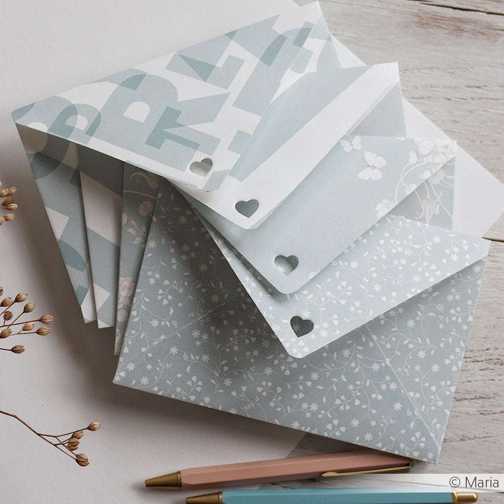 Stapel pastellfarben gemusterter Briefumschläge aus Alexandra Renke Designpapier mit Herzausschnitten, die neben zwei Stiften und getrockneten Blumen auf einer Holzfläche ausgestellt sind.