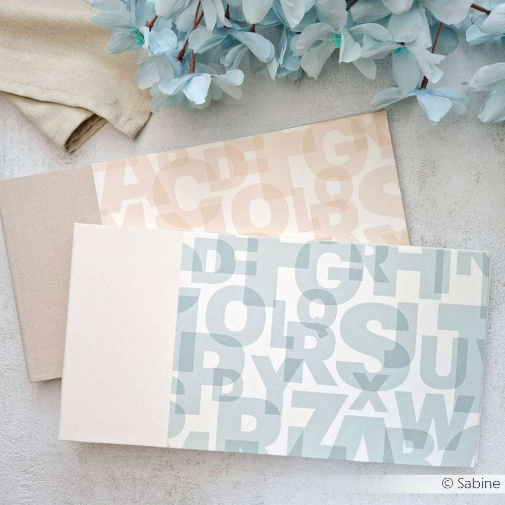 Zwei pastellfarbene Alphabet-Hefte von Alexandra Renke auf einer hellen Oberfläche mit blauen Blumen in der Ecke, perfekt für kreatives Buchbinden oder als einzigartiges Bucheinbandpapier.