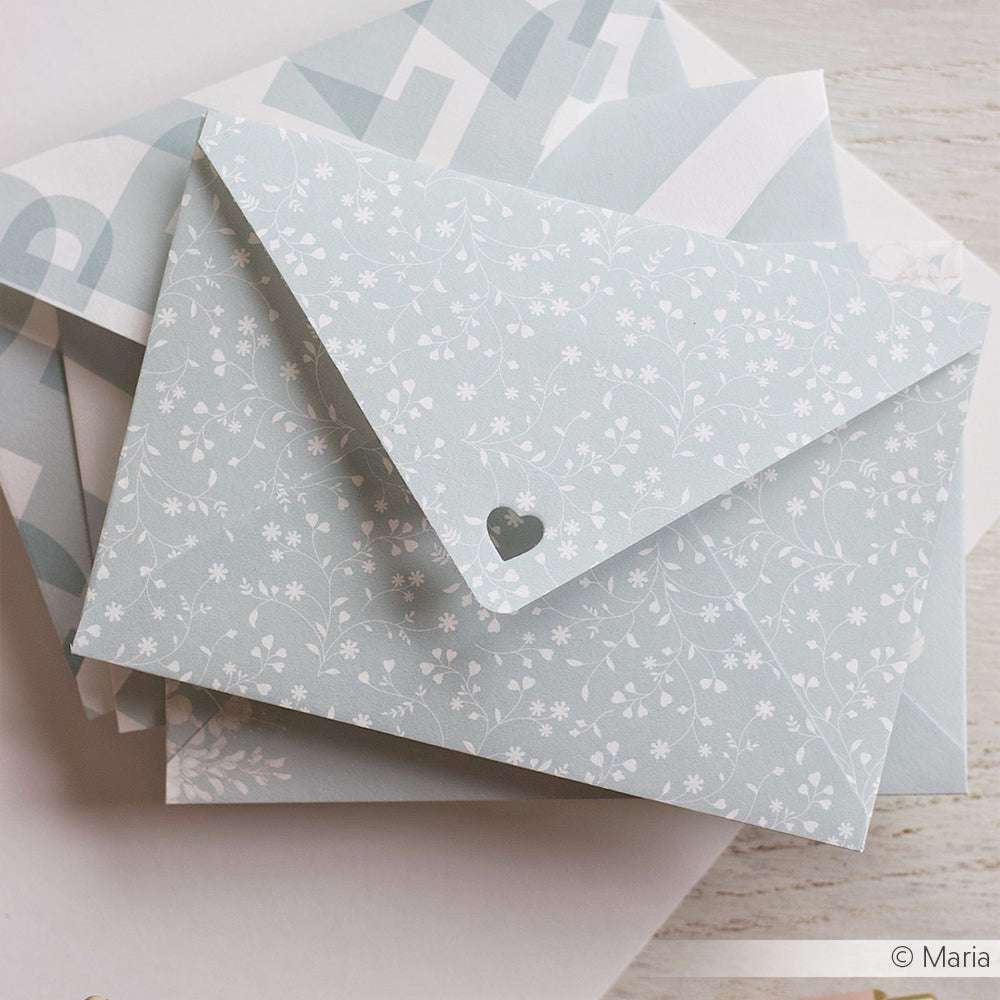 Ein pastellfarbener Blumenumschlag mit Herzausschnitt von Alexandra Renke ruht auf gemustertem Designpapier - ideal für kreatives Buchbinden oder als charmantes Bucheinbandpapier auf einer hellen Holzfläche.