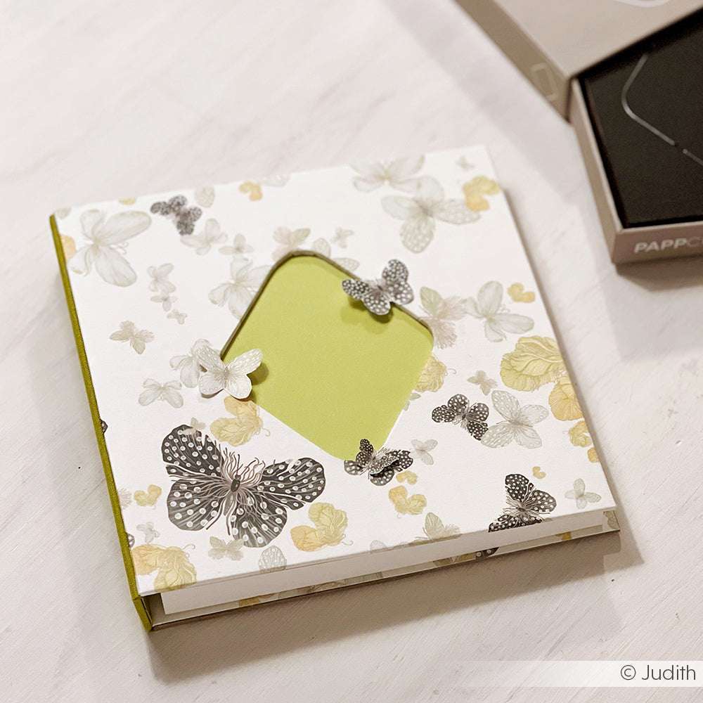 Quadratisches Buch von Alexandra Renke aus elegantem Designpapier mit Schmetterlings- und Blumenmotiv und einem ausgeschnittenen Fenster auf dem Einband, ausgestellt auf einer weißen Fläche.