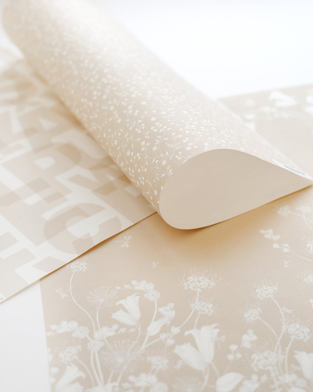 Eine Rolle beigefarbener Tapete mit weißen floralen und geometrischen Mustern, die teilweise auf einer ebenen Fläche ausgerollt wird.