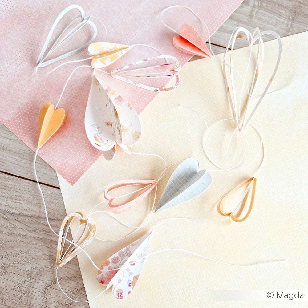 Die pastellfarbenen Papierherzgirlanden von Alexandra Renke sind auf hellrosa und cremefarbenen Blättern auf einer Holzunterlage angeordnet.