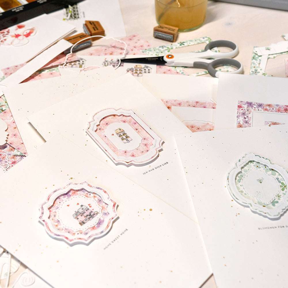 Alexandra Renke handgefertigte Grußkarten mit floralen Motiven sowie Scheren und Bastelutensilien auf einem weißen Tisch - ideal für kreative Sammlungen oder charmante Kartenbündel.